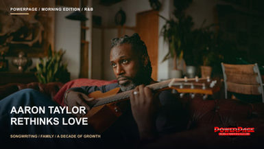 Aaron Taylor holds an acoustic guitar while reclining on a brown leather sofa.