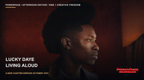 Lucky Daye stands in profile against a warm red background, wearing a brown jacket and gold necklaces.
