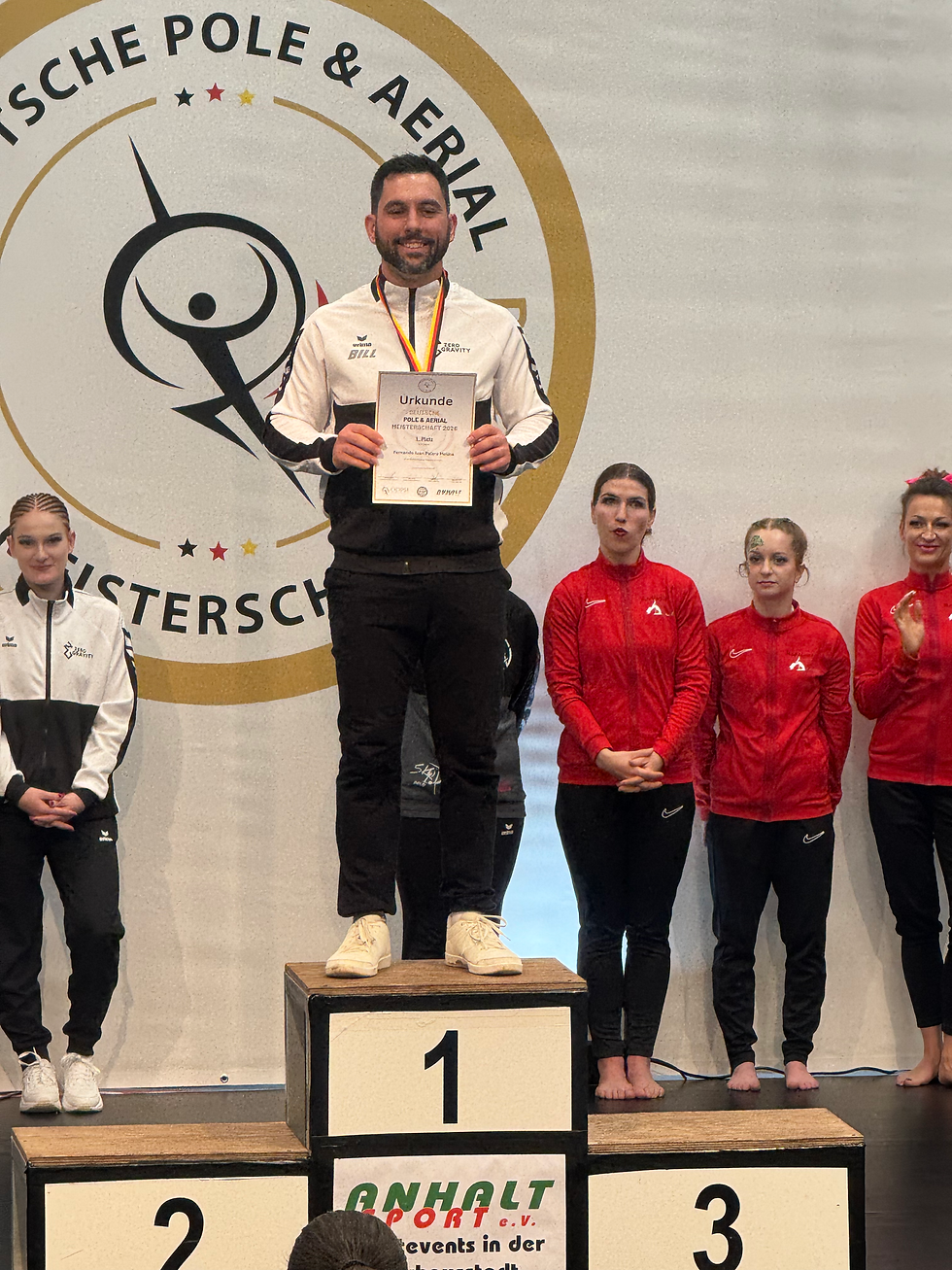 Man stands on podium holding a certificate, smiling, surrounded by others in athletic wear. "DEUTSCHE POLE & AERIAL" text in background.
