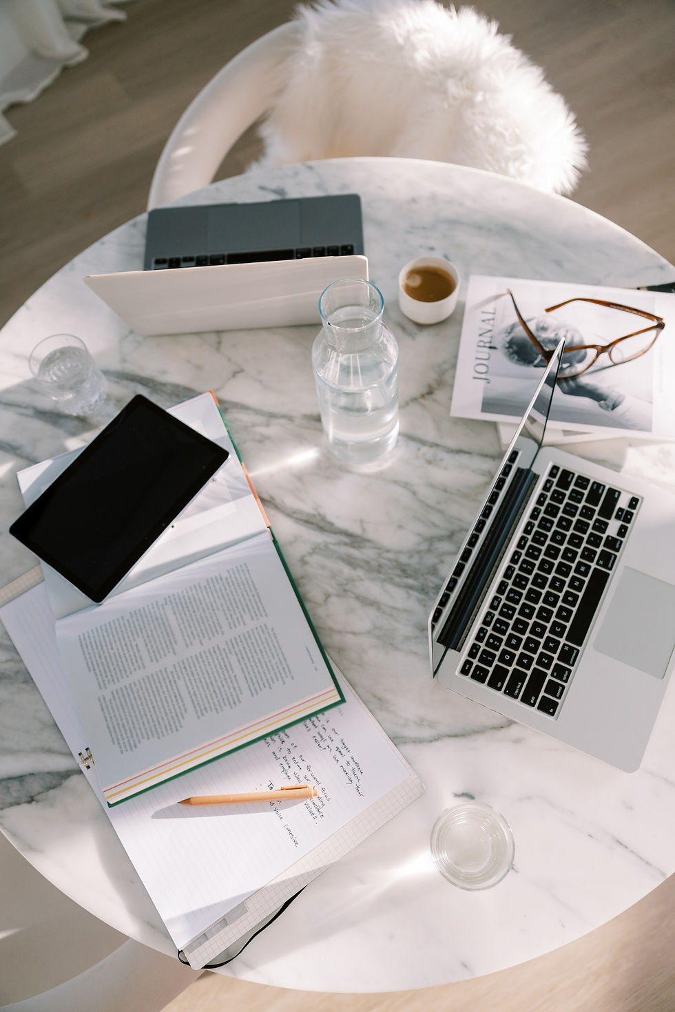 Marble table with laptops, tablet, open notebook, pencil, water bottle, coffee cup, glasses, and a journal. Bright, serene setting.