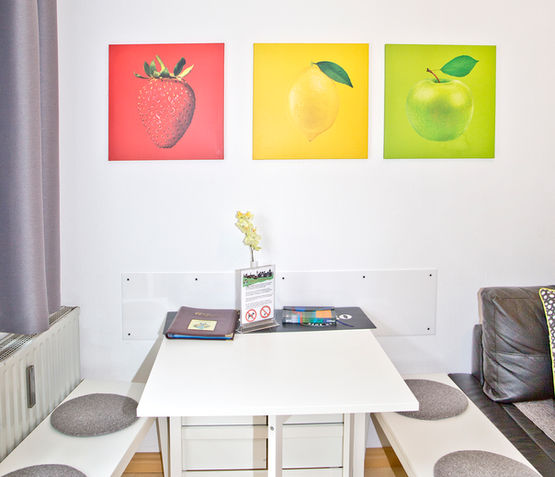 Gemütliche Sitzecke im Apartment Nils der Steinhauser's Ferienwohnungen Augsburg mit weißem Tisch, Sitzbänken, grauen Kissen und bunten Frucht-Wandbildern.