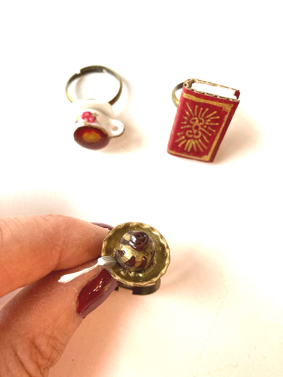 Thumbnail: main avec la bague cake miniature au chocolat et en arriere plan les deux autres bagues