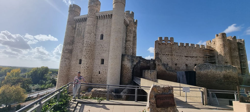 Persona junto a un castillo de piedra medieval bajo cielo azul, con torres y murallas; vista turística y soleada.