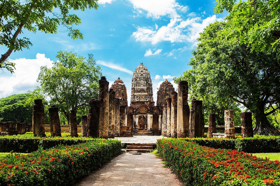 Ruinas de un templo antiguo entre árboles y flores rojas, con torre central de piedra bajo cielo azul y nubes.