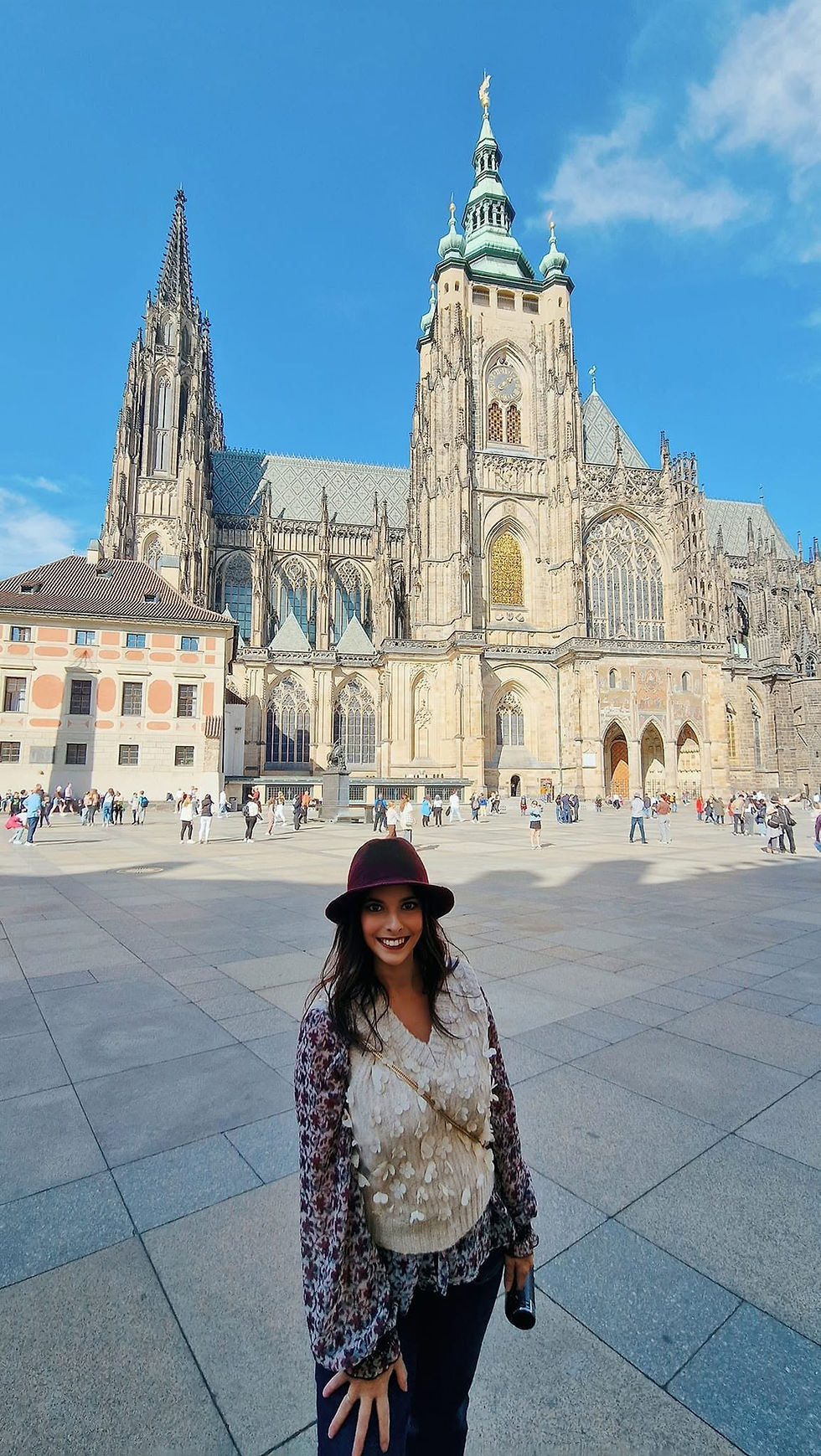 Mujer sonriente con sombrero en una plaza, frente a una catedral gótica; cielo azul y turistas al fondo.