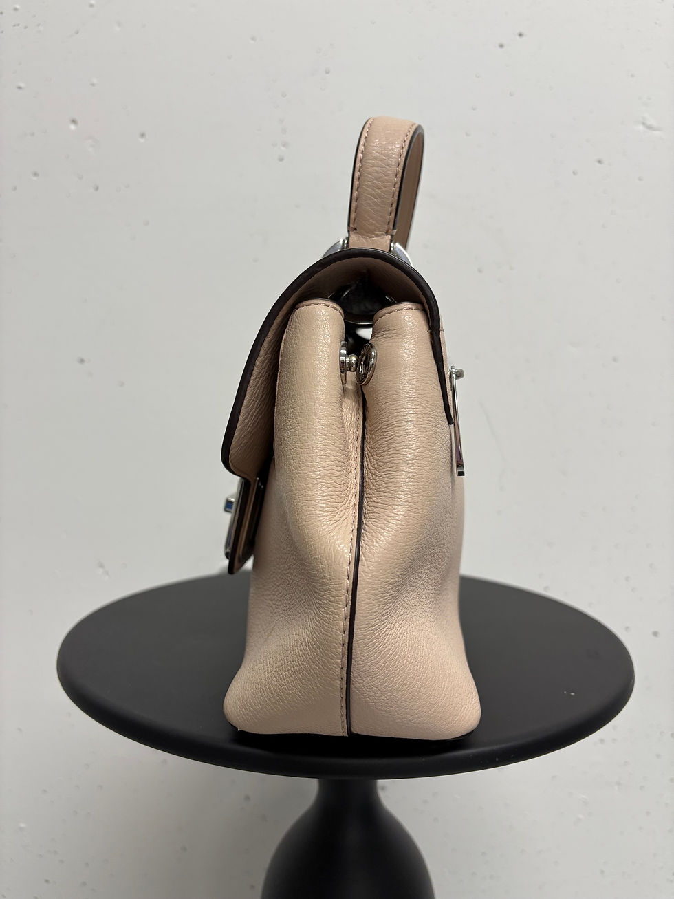 Beigefarbene Handtasche, seitliche Ansicht, mit Henkel und Verschluss auf einem kleinen Tisch.