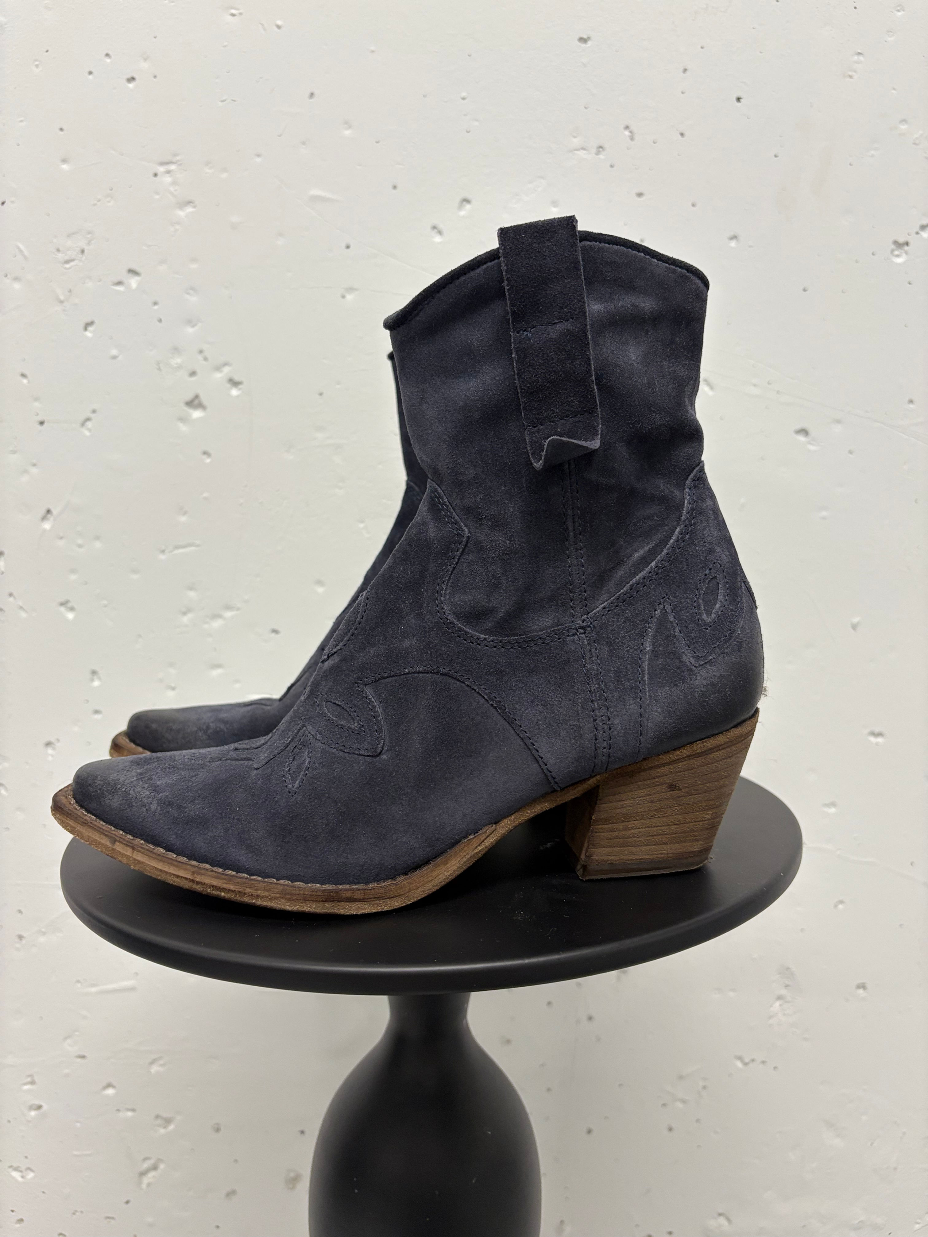 Dunkelblaue Wildleder-Cowboystiefel auf einem schwarzen Tisch vor weißem Hintergrund.