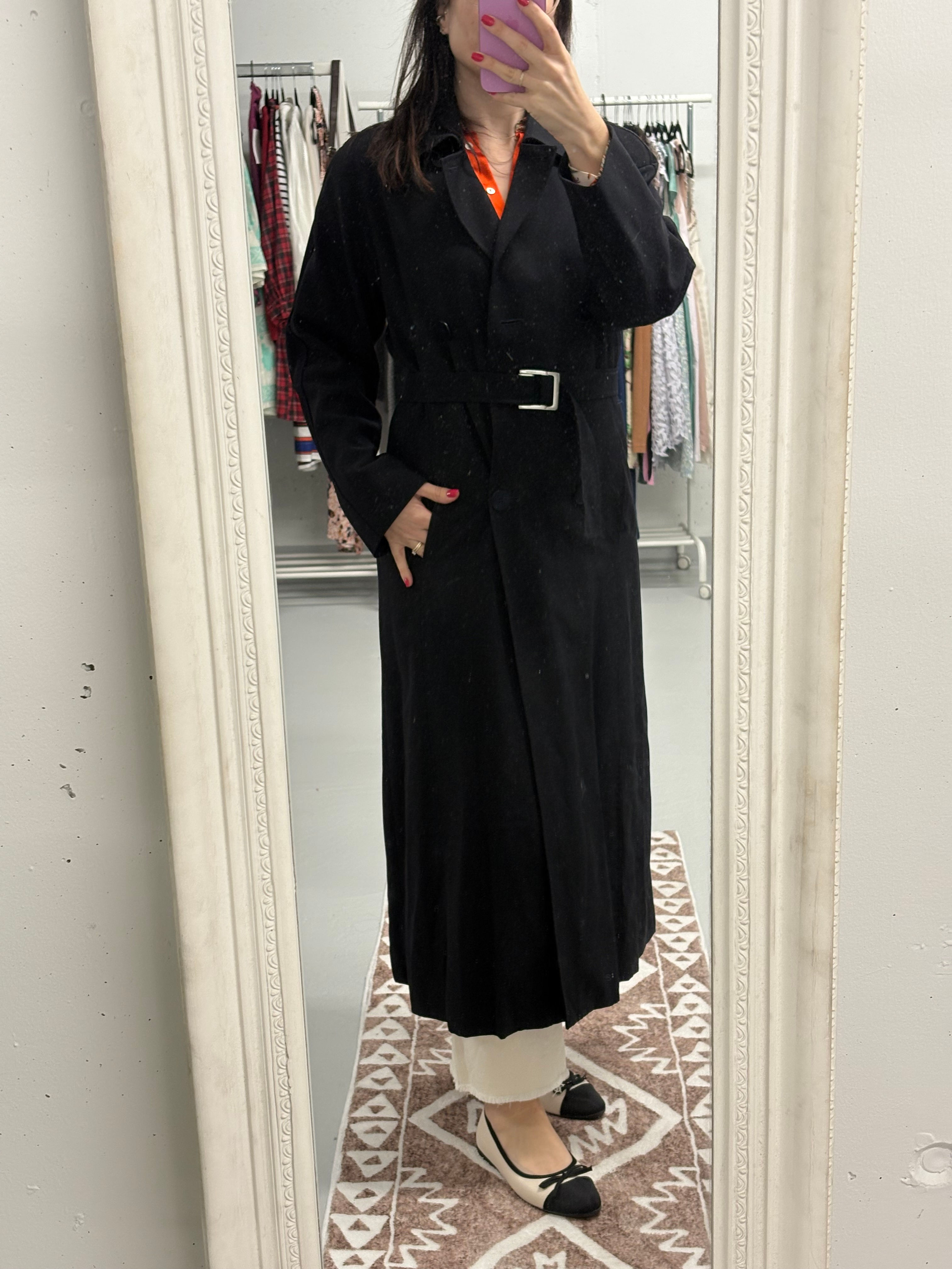 Frau trägt langen schwarzen Mantel mit Gürtel vor einem Spiegel. Kleidung im Hintergrund.