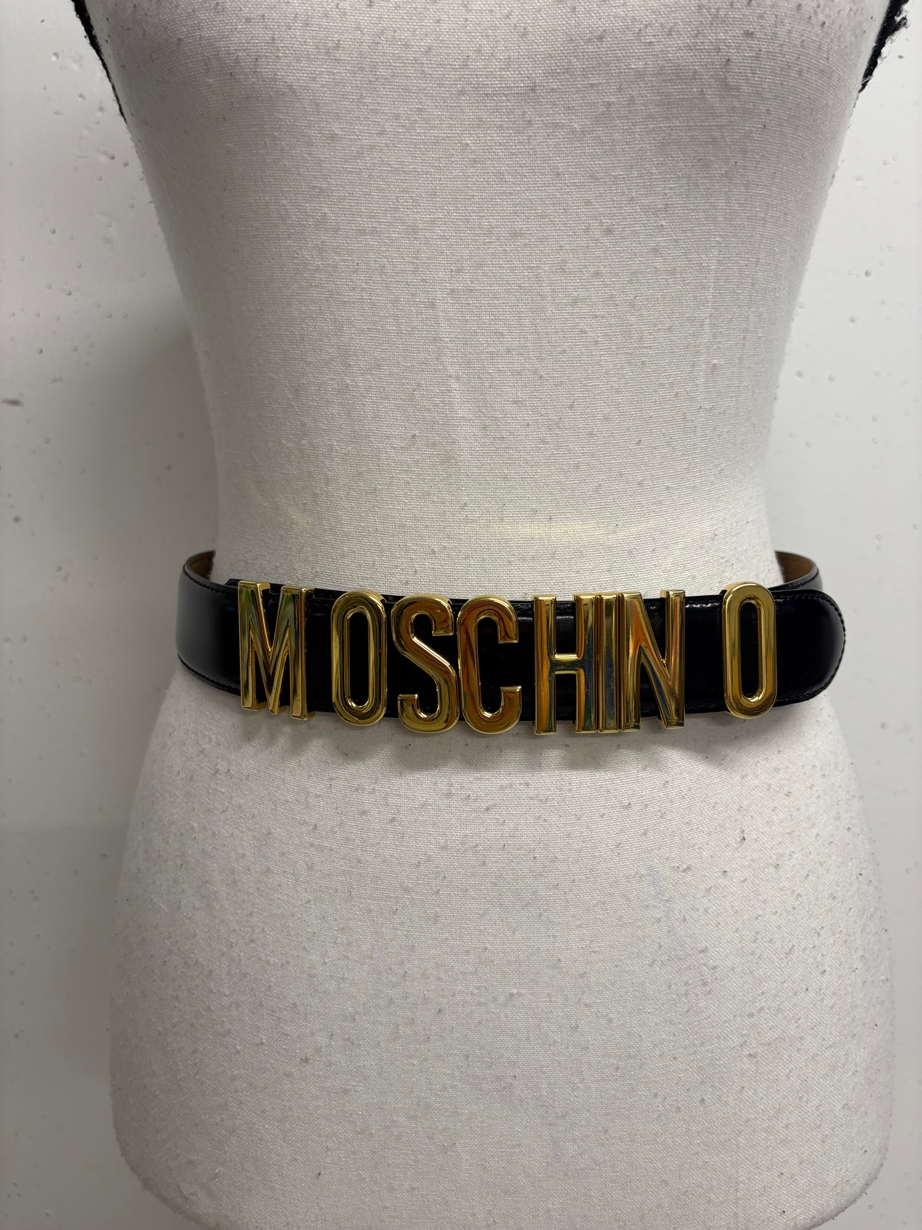 Schwarzer Moschino-Gürtel mit goldenen Buchstaben auf einem weißen Schaufensterpuppenkörper.