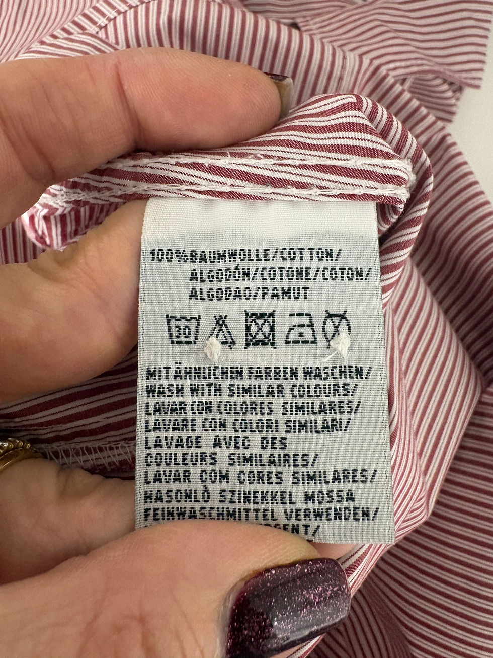 Miniaturbild: Pflegeetikett mit Text: Baumwolle, Waschhinweise, verschiedene Sprachen, rotes Hemd, Detailaufnahme.