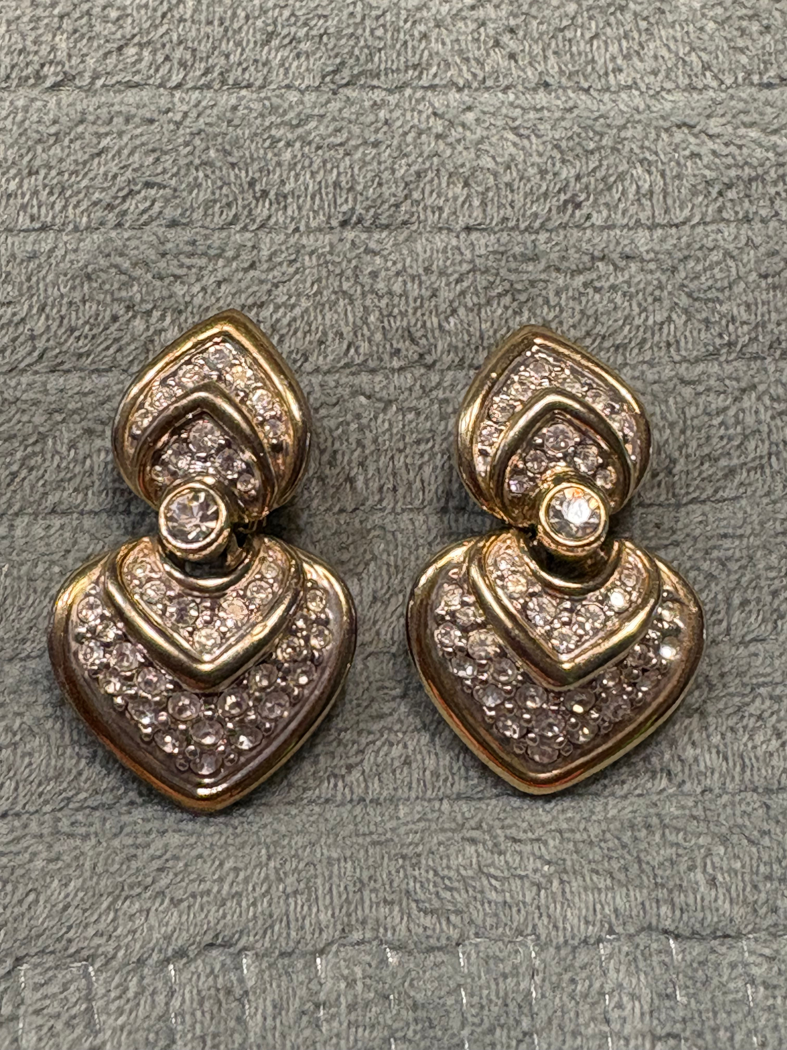 Paar goldfarbene Ohrringe mit funkelnden Kristallen vor grauem Hintergrund.