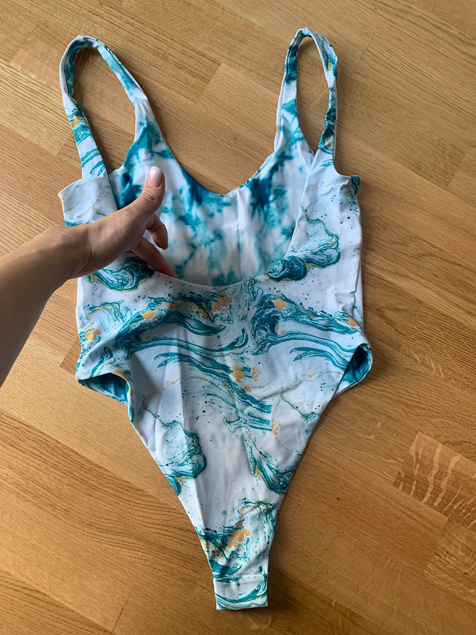 Miniaturbild: One-piece swimsuit, white and turquoise marble pattern, hand touching fabric on wood floor.