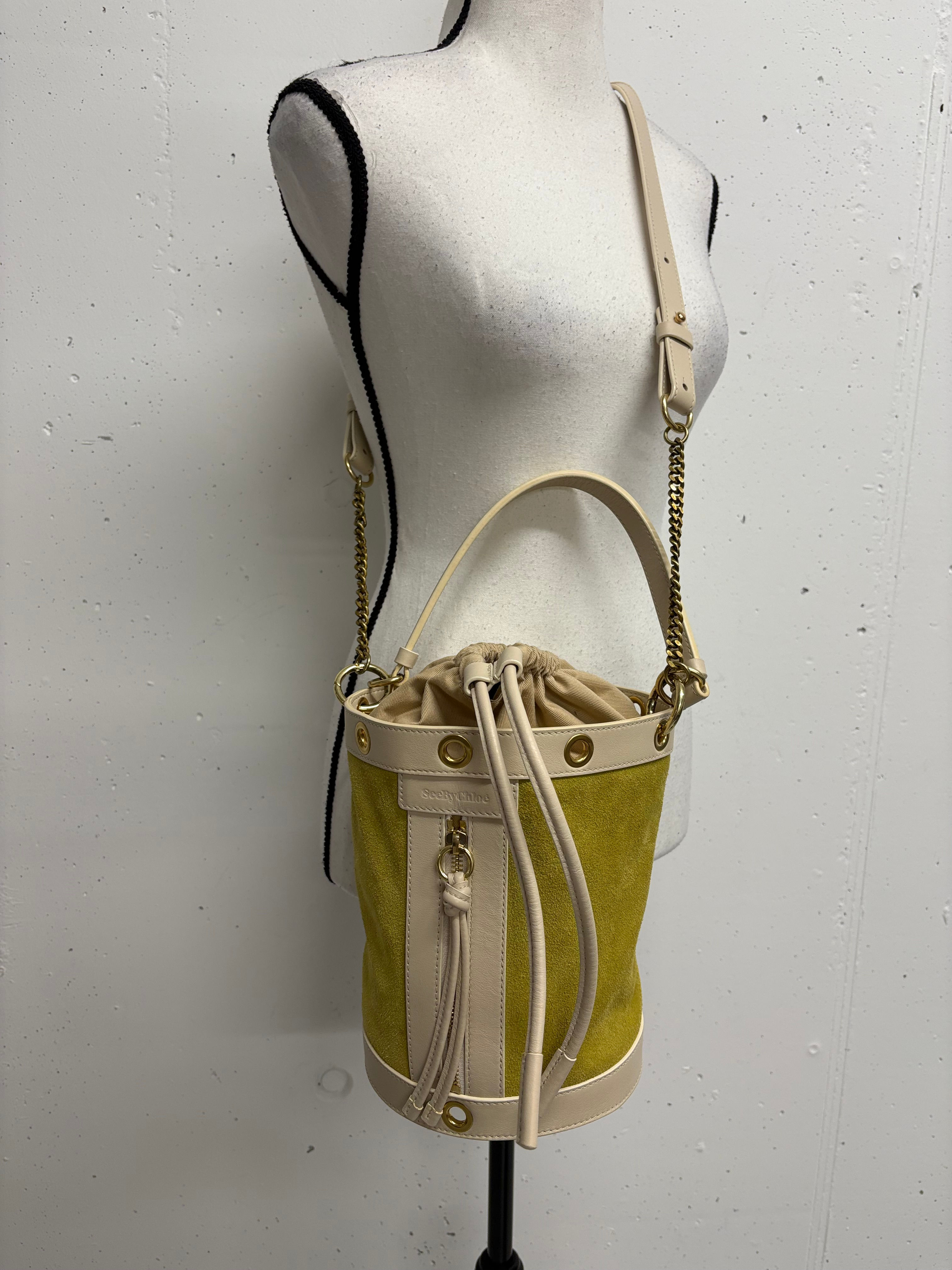 Gelbe Wildleder-Eimertasche mit Kordelzug Detail und Schulterriemen auf Schaufensterpuppe.