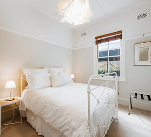 Queen bed with crisp white linen in bedroom with lamp, window, and mirror. White walls.