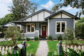 Bilawi Bungalow front view, classic 1930s Californian bungalow with dusty blue stucco finish, red front door, white detailing and window frames, picket fence, front lawn with elegant shrubs and trees