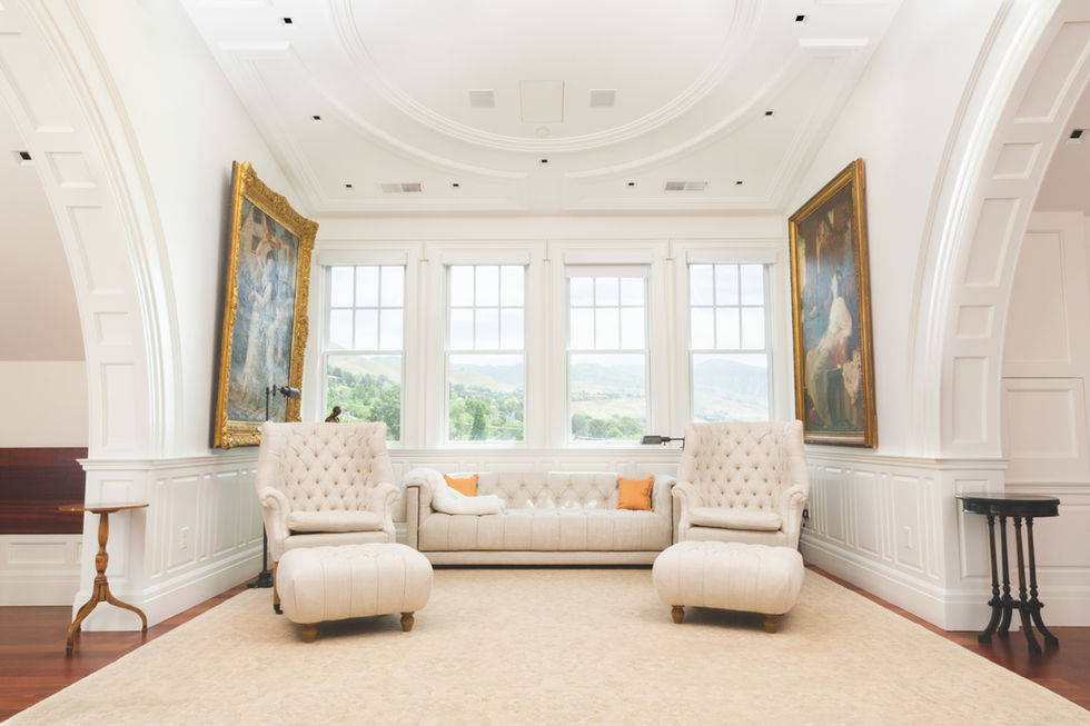 Bright sunroom with two cream armchairs, a matching sofa, large windows, and gold-framed art.