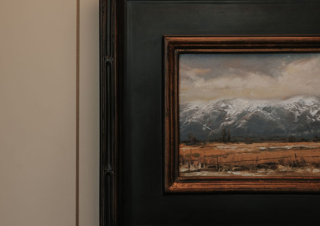 Framed landscape painting of snowy mountains and a field, mounted on a cream-colored paneled wall.