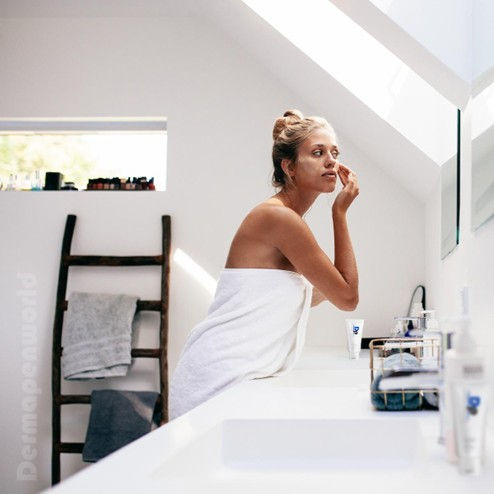 Woman standing over her bathroom sink in a towel, applying DP cream as aftercare treatment for her microneedling procedure.