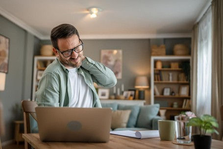 One man sitting at a kitchen table doing work on his laptop, suffering from a neck strain due to prolonged desk work.