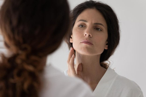 Woman with long, dark hair looking at her face in the mirror, wearing a white bathrobe and holding her hand to her lower jawline.