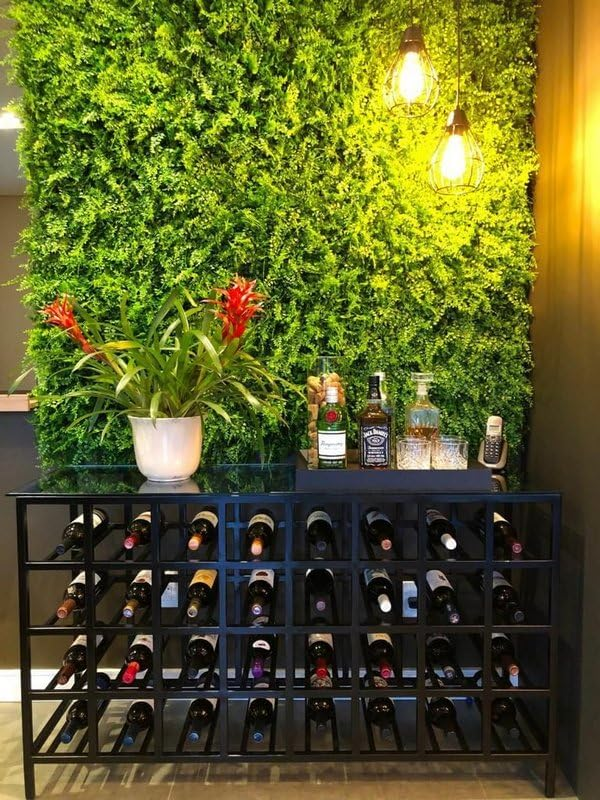Cantinho gourmet com adega de metal preta para vinhos, bancada com garrafas de bebidas e vaso de planta com flores vermelhas. Ao fundo, um exuberante jardim vertical verde e duas luminárias pendentes com lâmpadas de filamento, criando um ambiente aconchegante e sofisticado.