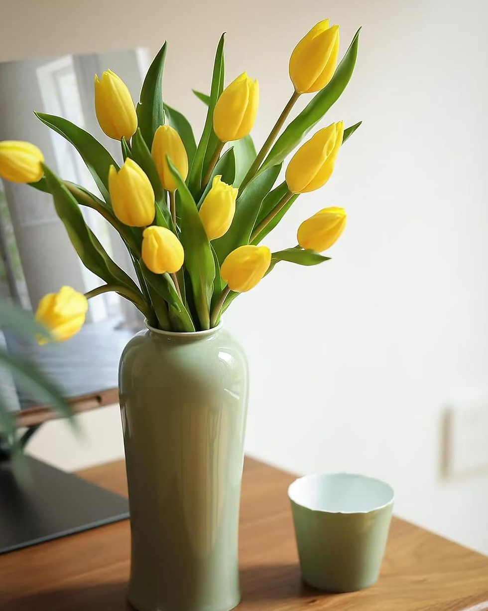 Vaso de cerâmica verde com arranjo de tulipas amarelas, apoiado sobre uma mesa de madeira. Ao lado, há um pequeno copo na mesma tonalidade do vaso. Ao fundo, um ambiente iluminado e minimalista, trazendo sensação de frescor e elegância.