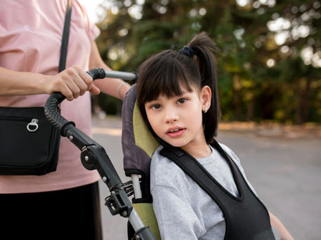 A child in an adaptive stroller is looking at the viewer. A person is standing behind the stroller.