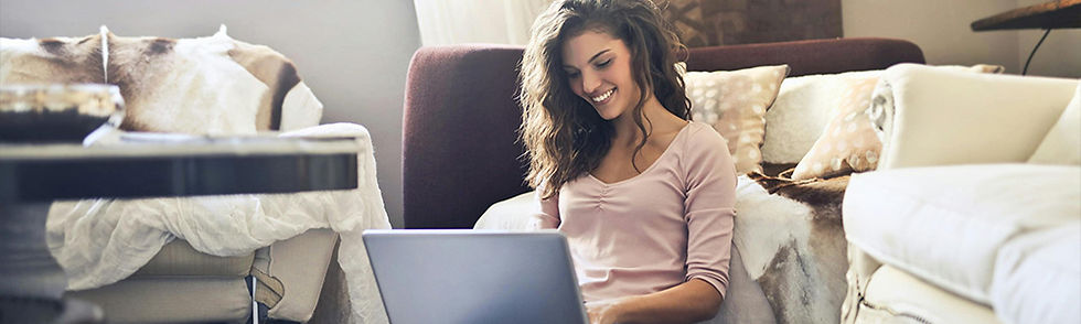Female content creator smiling while using a laptop, seated on a cozy couch with pillows and blankets in a warmly lit living room, creating a relaxed mood.