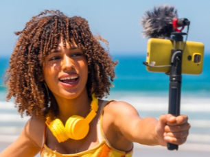 Content creator with curly hair taking a selfie video with a phone on a tripod at the beach, wearing yellow headphones. Ocean and sky in the background.