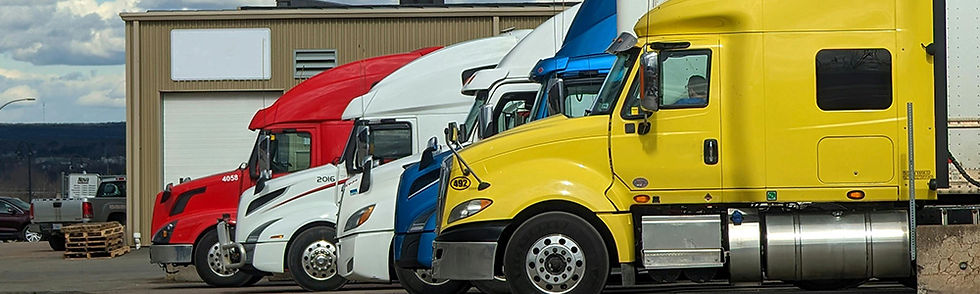 Four colorful semi-trucks (red, white, blue, yellow) parked side by side at an industrial area under a cloudy sky. Building in the background.