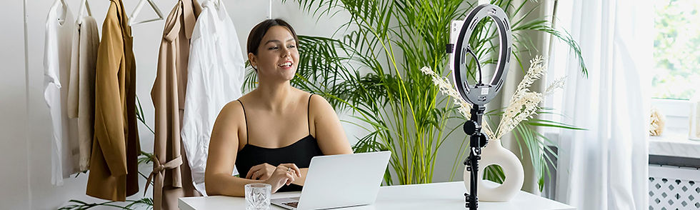Female content creator in black top sits at a desk with a laptop, smiling in a bright room. Ring light and clothes rack in the background, plants nearby.