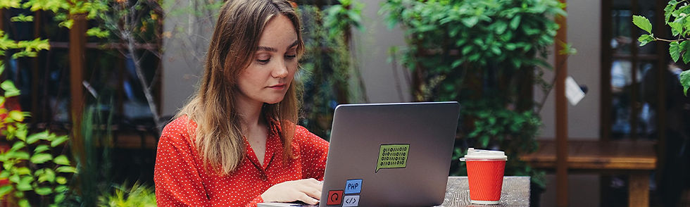 Woman in red shirt works on a laptop with green sticker, beside a red coffee cup. Setting is an outdoor café with greenery.