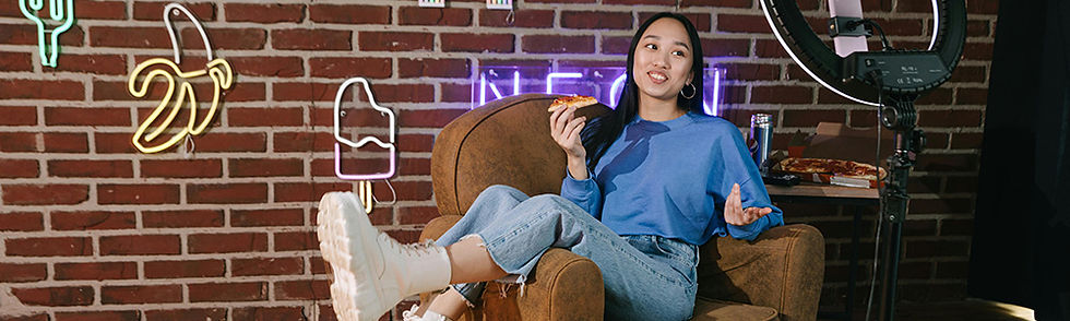 Woman in a blue shirt sits on a brown chair, eating pizza. Neon signs of a banana and ice cream are on the brick wall background.
