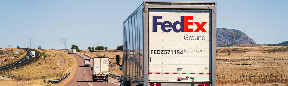 A FedEx truck drives down a rural highway under a clear blue sky. Other vehicles are visible ahead. Text on truck reads "FedEx Ground".