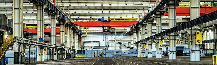 Spacious industrial warehouse interior with red overhead crane systems, white columns, and yellow signs. Floor marked with lines; no people visible.