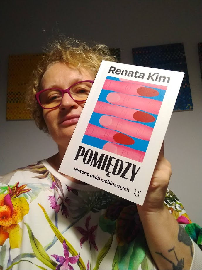 Kobieta patrząc w obiektyw aparatu trzyma książkę Renaty Kim pod tytułem Pomiędzy. Historie osób niebinarnych. W tle kolorowe obrazy