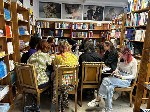 Grupa 10 młodych osób siedzi przy stole, skupiona na pracach manualnych z tkaniną. W tle regały z książkami.