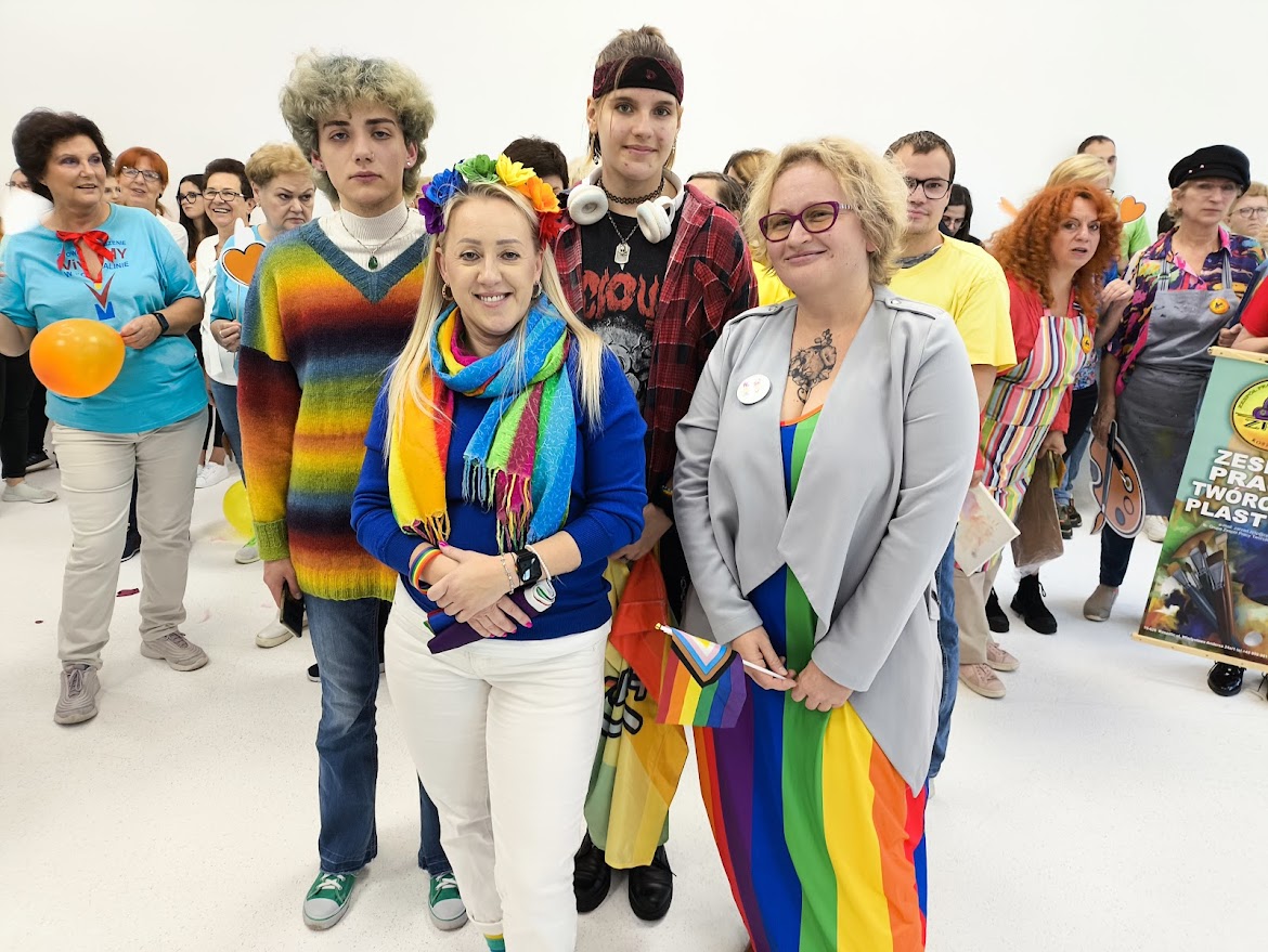 grupa osób w tęczowych strojach uśmiecha się do zdjęcia, w tle tłum społeczników