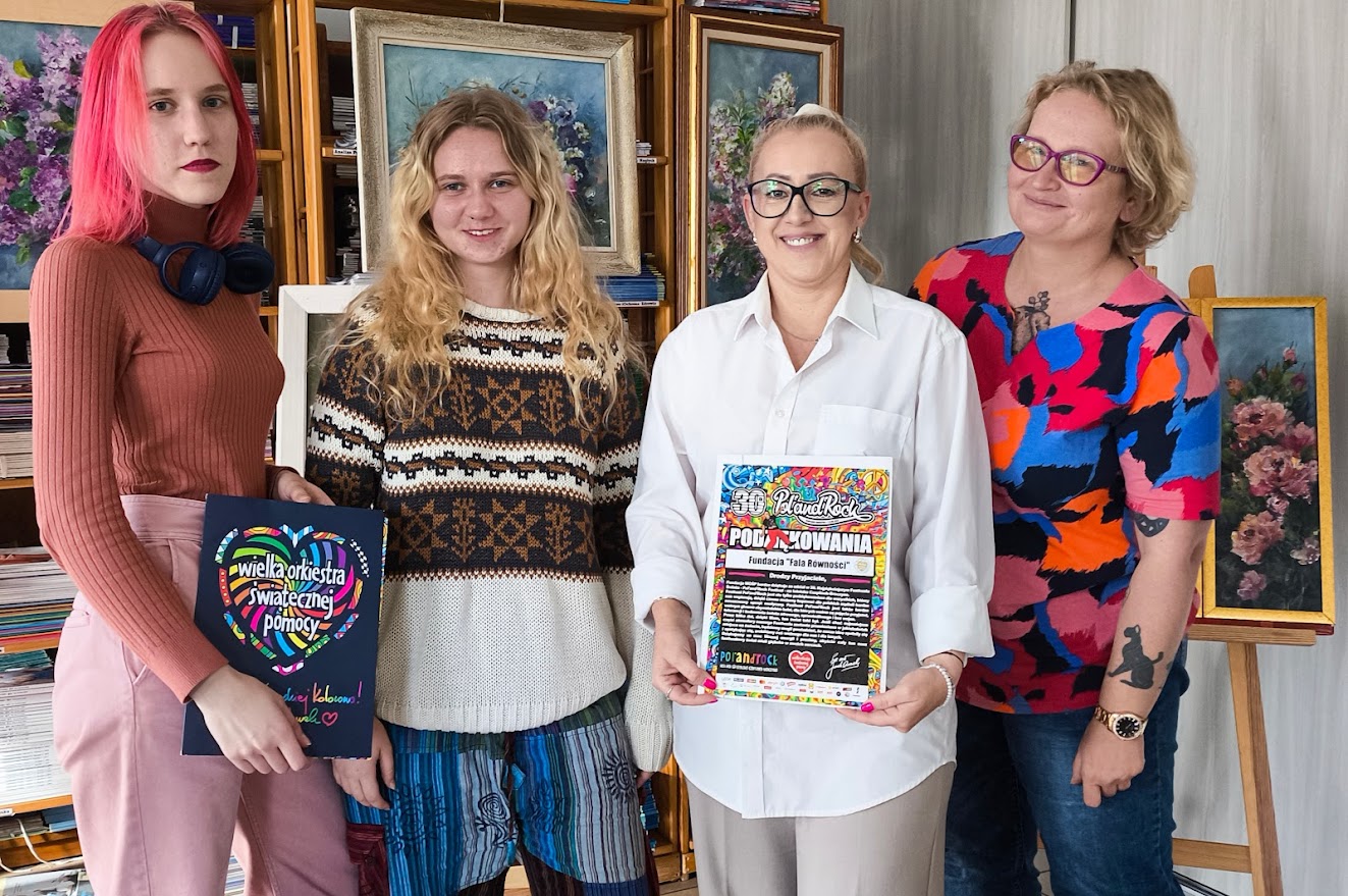 4 kobiety usmiechają się do aparatu, trzymając dyplom - podziękowanie od Jurka Owsiaka za udział w festiwalu Pol'and'Rock, w tle obrazy z kwiatami
