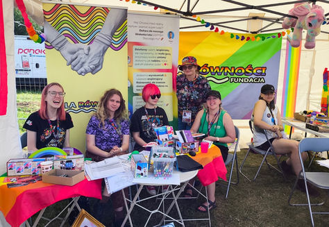 Grupa 6 osób w namiocie pozuje do zdjęcia. Wokół tęczowe flagi, rollupy i gadżety oraz stolik z wlepami i ulotkami dot. LGBT+