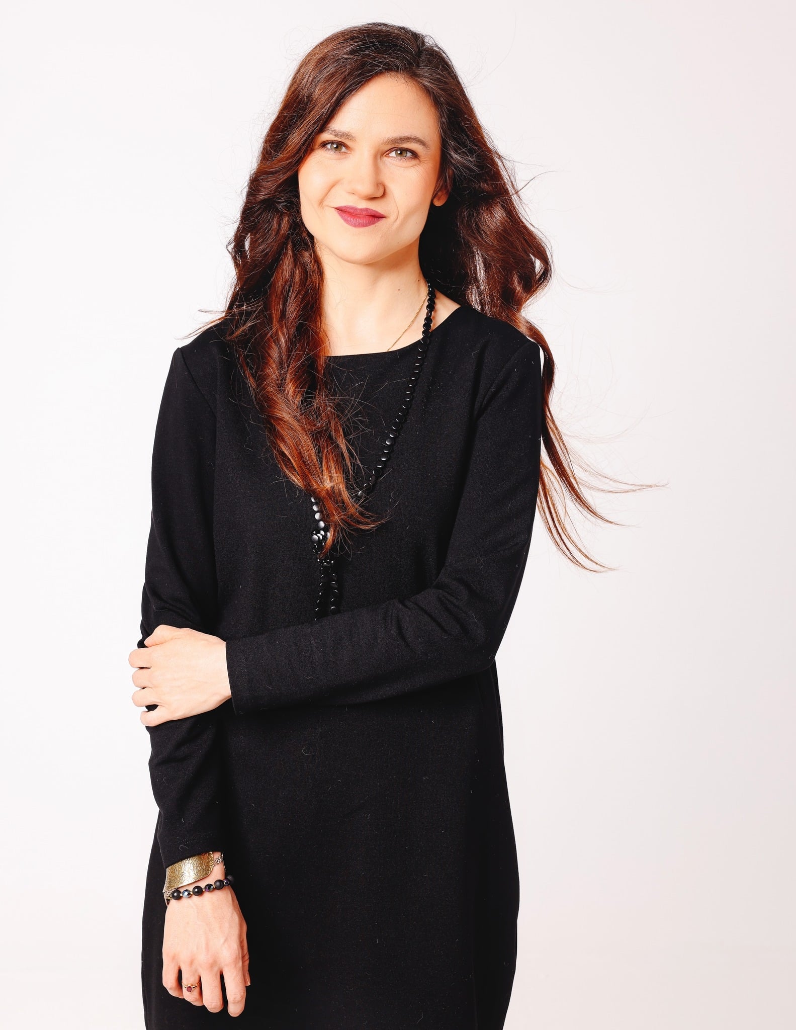młoda kobieta z rozpuszczonymi włosami, uśmiechnięta, w czarnej sukience stoi na tle białej ściany