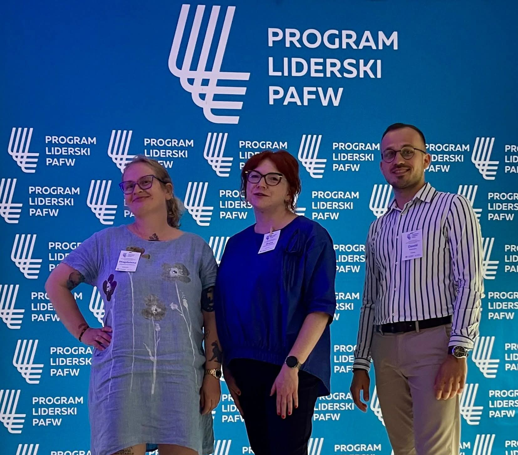 trzy uśmiechnięte osoby stoją na tle niebieskiej ścianki z napisem: Program Liderski PAFW