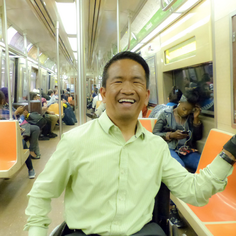A Asian American man sits on the subway smiling. He holds onto a pole for support. He is a wheelchair user, and wears a lime green shirt..