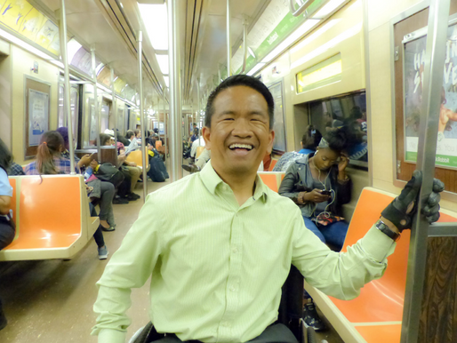 A Asian American man sits on the subway smiling. He holds onto a pole for support. He is a wheelchair user, and wears a lime green shirt..