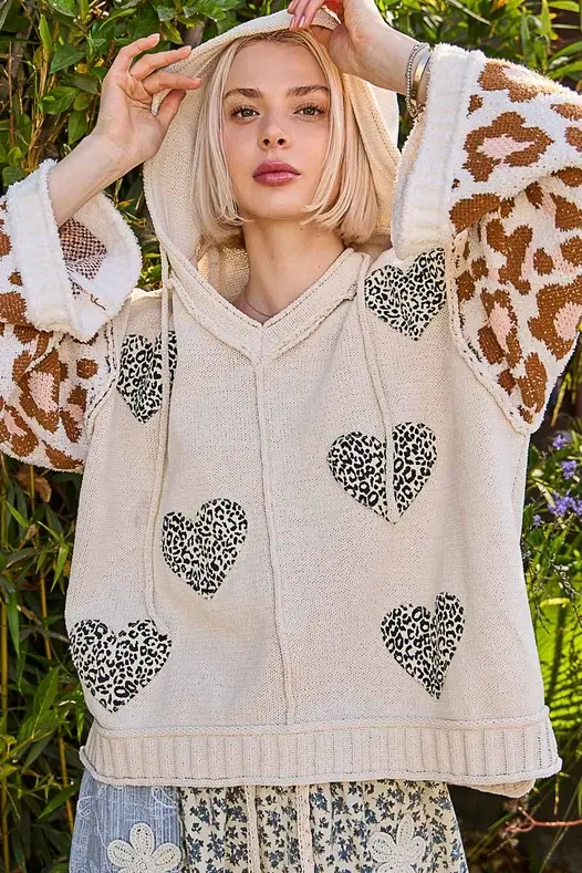 Thumbnail: Cream sweater with black heart patches and long giraffe-print sleeves, shown with the model’s hands on her head.