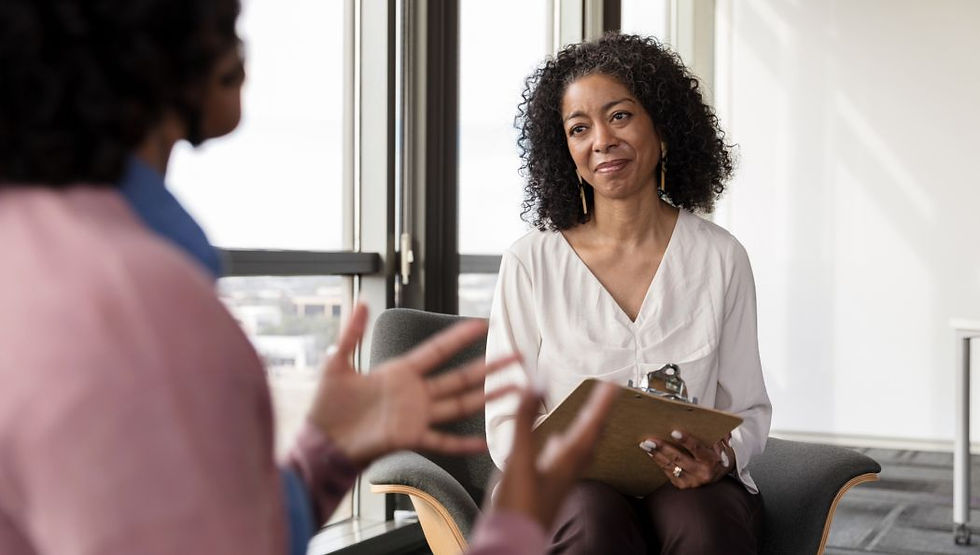 Woman listens with clipboard in bright office as blurred speaker gestures in foreground, suggesting a calm counseling session