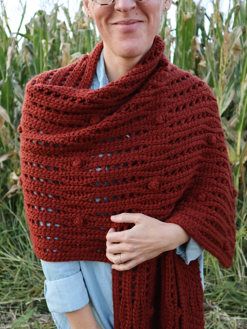 pinterest pin of a girl draped in a handmade copper colored crochet shawl featuring undulating details of ribbing, lace, and bobble stitches