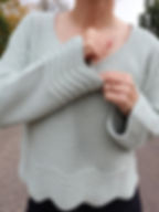 girl modeling handmade crochet sweater in the color light sage featuring a v-neck, long sleeves, and a unique ribbed wavy bottom hem and cuffs.