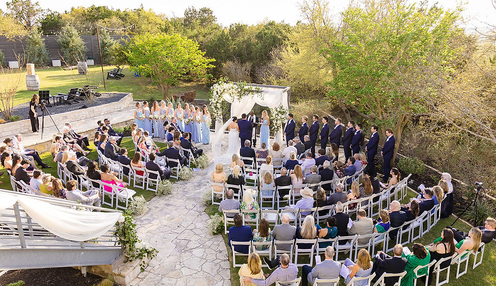 A stunning outdoor wedding ceremony at The Terrace Club in Dripping Springs, Texas, surrounded by lush Hill Country greenery and golden afternoon light. Guests look on as the couple exchanges vows beneath a romantic floral arch draped in soft white fabric. With natural stone pathways, manicured lawns, and breathtaking views, this space captures the perfect balance of elegance and Texas Hill Country charm that makes The Terrace Club one of the most sought-after wedding venues near Austin.