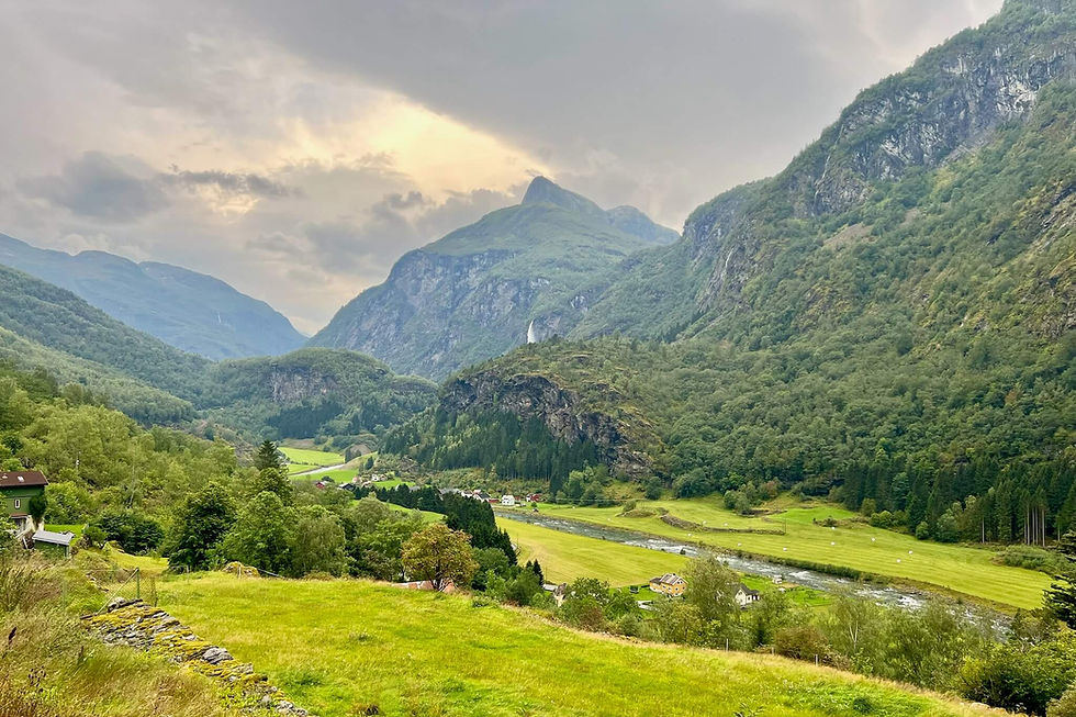 Green mountain valley with river, scattered houses, and steep cliffs under cloudy sunset sky, serene rural scene in Norway. 